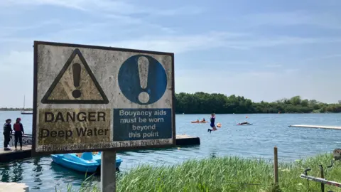 A sign in the foreground says "Danger deep water buoyancy aids must be worn beyond this point. In the background children in lifejackets can be seen standing on a long a wooden walkway towards a lake, with one person jumping into the water.