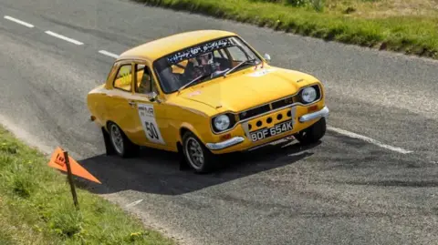 LUCAS CROYDON PHOTOGRAPHY A yellow classic rally car with the number 50 on the side. It is driving on a road with grass verges on either side.