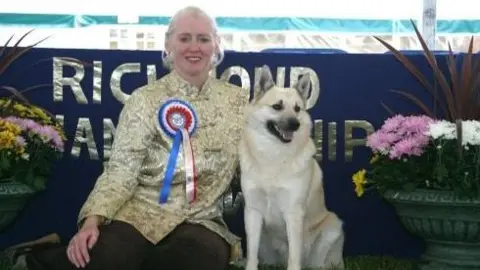 Di Sterling Mrs Sterling wearing a gold top with a red, white and blue rosette pinned. She has her blonde hair tied back in a ponytail and is sitting on the floor next to her Norwegian Buhund