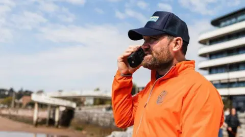 Will Reddaway Brendon Prince wearing a cap and an orange jacket, looking out to sea and holding a walkie talkie