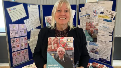 Head and shoulders shot of Chris Suich. She has blonde shoulder-length hair and is wearing a black top. She is holding one of the kit packs, which has a picture of her husband Bob on the front.
