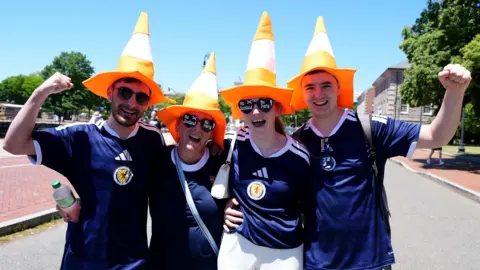 PA Media Four Scotland football fans wearing blue Scotland football shirts and orange traffic cone hats. They are cheering towards the camera.