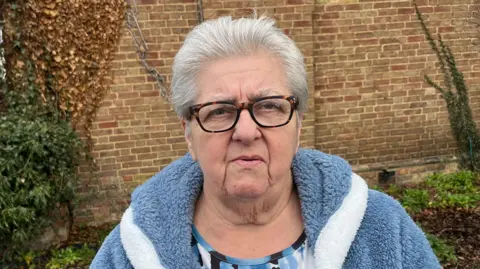 John Devine/BBC Anne Wells has short grey hair and is wearing brown and black leopard print framed glasses. She is wearing a blue, black and white patterned T-shirt and a blue coat. She is standing in front of a brick wall. 