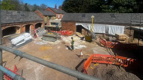 Building work under way at a stable block, showing a man in the middle, wearing a hard hat and hi-vis. The buildings are made of bricks, building items are in the middle, including red plastic barriers. Sheets cover some of the windows. 