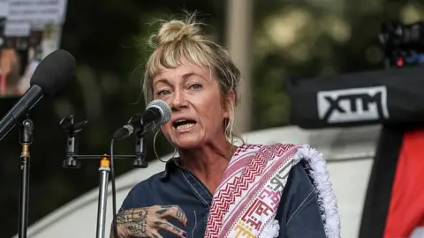 A blonde-haired woman with a keffiyah scarf over her shoulder speaks into a microphone