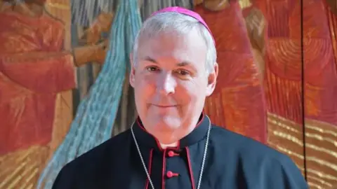 Diocese of Northampton Bishop of Northampton David James Oakley. He is wearing a black clerical gown and a pink skull cap.