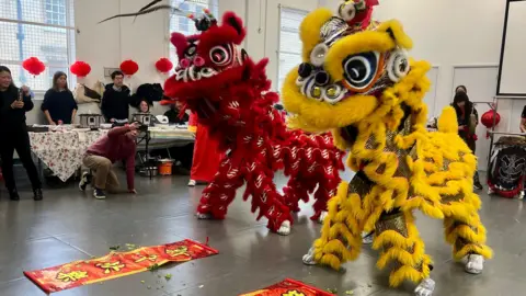 Teresa Lee Image shows a red and a yellow dragon costume, someone is acting as the head and someone acting as the feet. They are doing the traditional lion dance. There is a mat with writing in cantonese. There are people on the sidelines holding up their phones.