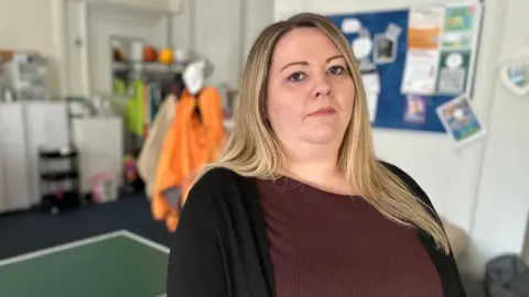 Carol Ann has long blonde hair and wears a maroon top and black cardigan