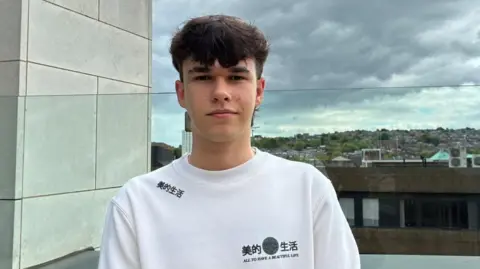 A young man with dark brown hair in a mullet style stands in front of view of Barnsley skyline. He wears a white jumper with black Chinese symbols on one should and a logo on the chest.
