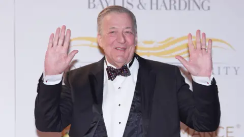PA Media Sir Stephen Fry in a dinner jacket with a colourful spotty bow tie and his hands in the air
