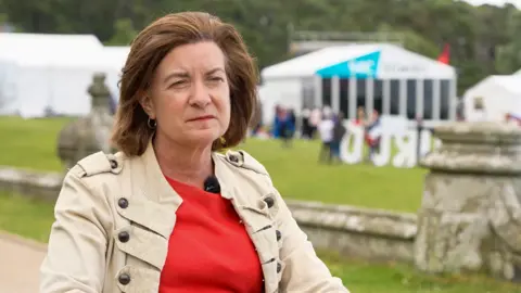 A woman with bobbed dark brown hair, wearing a red blouse and beige jacket and sat with a green field with white tents in it behind her.