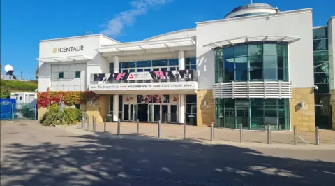 Google The entrance to the Centaur on a sunny day. The building is made from white concrete and yellow bricks.