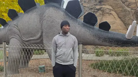 Family handout Eghosa stands, wearing a grey hooded top and black trousers, with a black hat, smiling slightly and with his hands in his pockets. Behind him is a large model dinosaur. 