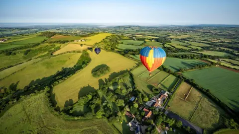PA Media A hot air balloon with its main body coloured red, yellow and blue, is seen flying over Bruton in Somerset, with multiple green fields and hedgerows below