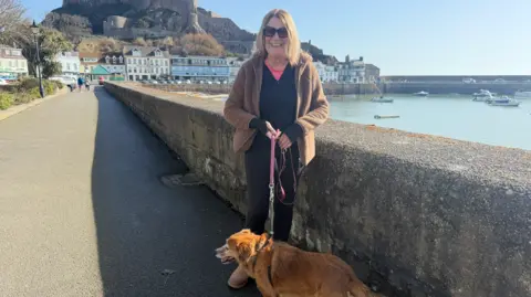A woman, in her 60's walking along Gorey seaside path holding a dog’s lead, with the dog standing beside her. A stone wall lines the path, and a harbour with boats is visible to the right. Houses and a large hilltop castle, Gorey Castle, appear in the background under a clear sky.