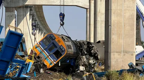 A damaged train lying on its side surrounded by twisted metal with overhead rail tracks in the background