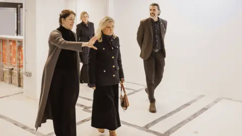 The Lighthouse A woman with dark hair talks to a blonde woman (Duchess of Edinburgh) with a man and a woman in the background inside an empty room.