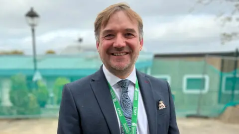 Stuart Woodward/BBC Martin Hawrylak wears a navy blue suit with a white shirt, blue patterned tie and a green lanyard around his neck. He has short brown hair and a stubbly beard, and is smiling at the camera. He stands in front of a building which has fencing around it.
