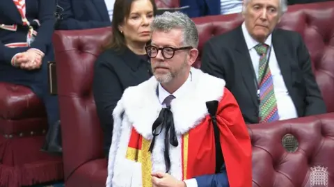 UK Parliament/PA Lord Doyle wears a red robe with a white collar over his suit. He is stood in front of the red benches of the House of Lords.
