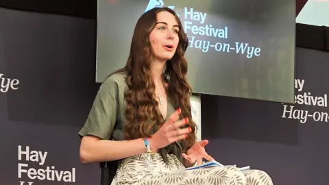 Bethany Handley Bethany on stage at Hay Festival. She wears a cream dress with a leaf pattern on it and a khaki short sleeved tie top. There is Hay Festival branding on the stage behind her. She is mid-speech looking at the audience to the right of the photo.