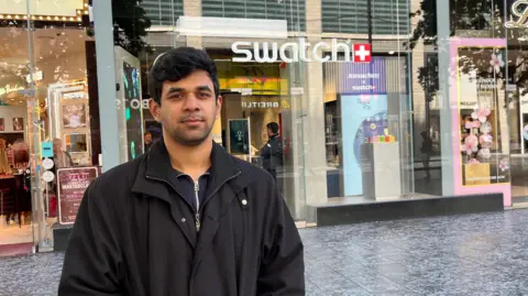 Jonny Humphries/BBC A 21-year-old man with black hair styled into a quiff, light stubble and a black overcoat looks into the camera outside a a glass-fronted shop with the Swatch logo above the door in illuminated white lettering.