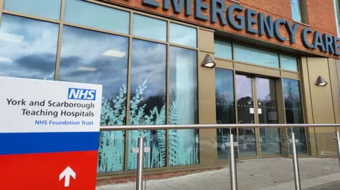 The exterior of a hospital building, with large glass windows and doors, and a sign that reads York and Scarborough Teaching Hospitals.