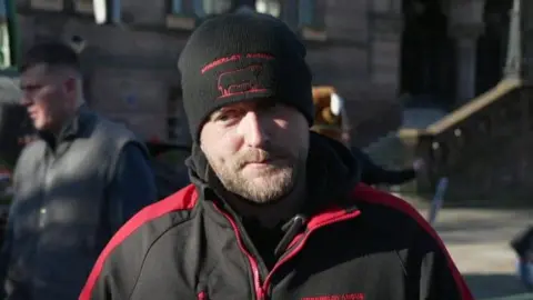 Ian Norbury has a short beard and moustache and is wearing a black hat and a black and red fleece with his family business' logo on it. The hat has a thin red outline of a farm animal on the front.