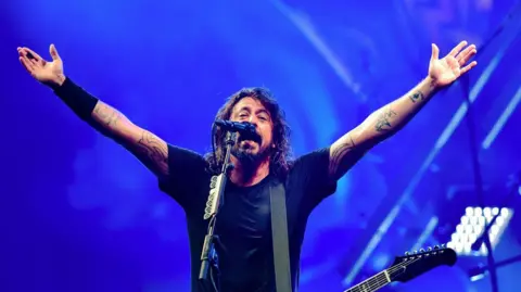 Dave Grohl, who is wearing a black t-shirt and has a beard with long brown hair, stands on stage with his arms stretched outwards. A guitar hangs from a strap over his shoulder while he faces a microphone. 