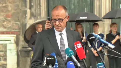 German chancellor in a suit speaking into microphones.