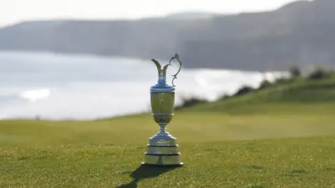 PA Media The silver claret jug sits on grass at Royal Portush Golf Club. You can see the headland and the sea in the background.