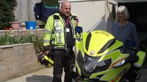 Russell Curwen is standing on a drive with an unnamed woman. He is wearing fluorescent emergency service clothing which has reflective stripes and there is a blood service motorbike in fluorescent yellow in front of him. 