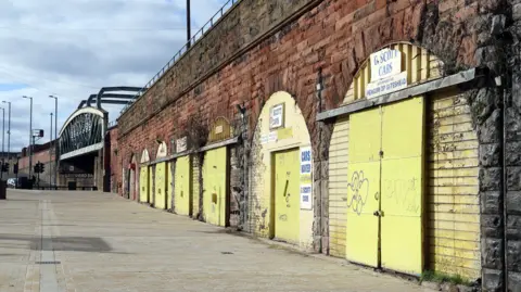 Gateshead Council Exterior view of a series of railway arches with old, fading yellow paint on the doors. There is a steel bridge in the distance.