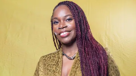 Jacqueline Shaw has long, dark purple braided hair. She is wearing a necklace and a light khaki patterned coloured top. She is standing in front of a light khaki background.