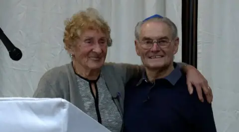 Susan Pollock and Tomi Reichental are standing side-by-side. Susan has curly red hair and is wearing a grey cardigan and top. Tomi Reichental is wearing a blue yamaka, a brimless, usually cloth skullcap traditionally worn by Jewish men, and a black sweater and navy shirt. 