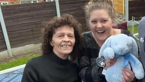 An older woman, two young women and a young man stand smiling at the camera as the young woman on the right takes a "selfie". The other young woman is holding a blue stuffed rabbit. 