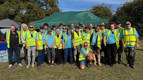 A large group of people wearing high-vis vests are gathered for a photo. They are stood in front of a green gazebo in a field. 
