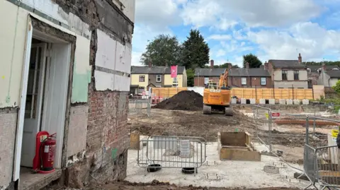 A building site with a brick building and large excavated area with fencing and a digger and terraced housing in the distance.