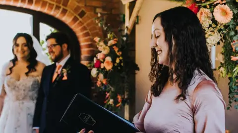 Sophie Ann Photography Zadie Ward, in a pink dress, reads from a black folder at a wedding