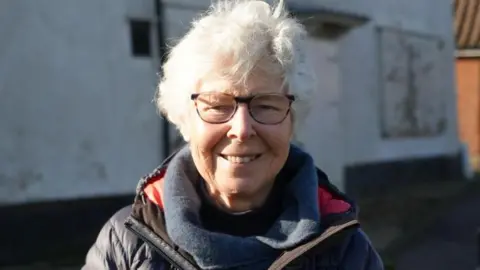 Shaun Whitmore/BBC Jill Halliday has white curly short hair. She has a navy wool scarf and a navy padded jacket. She is standing against a boarded-up building.