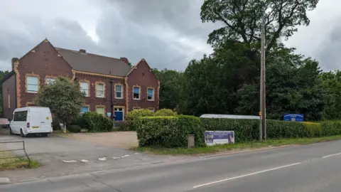 The school building set back from the roadside. There is a white van parked in the driveway and a banner and a blue sign outside. 
