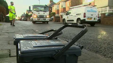 Two black plastic food caddies - about knee high - sit on a pavement in a Newcastle suburb. A council waste worker clad in a high-vis t-shirt and shorts strolls towards them in the background alongside a white waste removal van with headlights shining forward in the morning sun. 