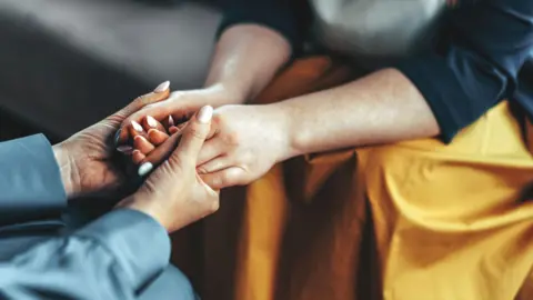 Getty Images Two people reach out towards each other and hold hands. They both have manicured nails. One person is wearing a light blue sleeved top, the other a black top with a yellow skirt.