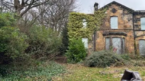 Front view of old stone building with windows boarded up and discarded furniture in the garden