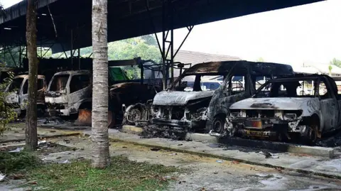 Five burned out vehicles are lined up under a tin roof structure