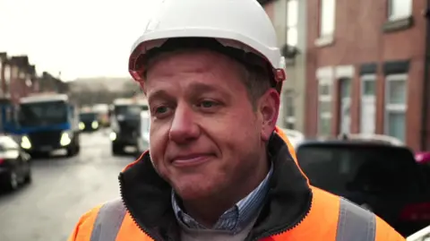 A man in a white hard hat wearing a blue shirt, grey jumper and orange high-vis jacket looks away from the camera. He is standing in a terraced street with construction vehicles behind him.