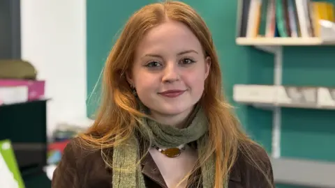 Olivia smiles at the camera. She has red hair and is wearing a brown jacket, green scarf and necklace. There are shelves of books behind her and a piano.