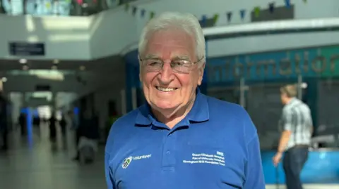 A man is a dark blue t-shirt with a "volunteer" sign on it. He is wearing glasses and smiling and the hospital is in the background. Another man is out of focus in the right of the picture. 