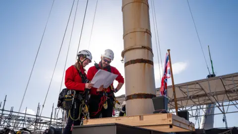 NMRN A man and a woman in climbing gear and helmets look at plans on the deck of the ship.