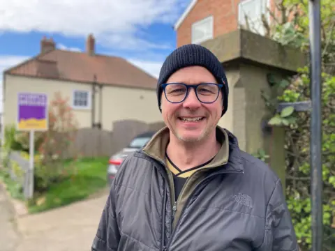 A man smiles as he stands outside the driveway of a smart-looking house. He is wearing a black "North Face" jacket, khaki fleece, blue-framed glasses and navy-blue woolly hat. A green garden, cream-coloured house and purple estate agent's sign can be seen in the background.