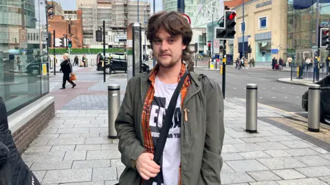 Ashley is stood in a city centre street with a grey/green jacket on over a checked shirt and white T'shirt with a black logo on. He has brown hair and a beard. Traffic lights and a junction can be seen in the background.
